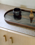 A dark oval serving tray with a warm brown wooden rim sits on a speckled stone countertop above light wood cabinets. On the tray are a black ceramic bowl and two small decorative bottles, with reflections visible on the glossy surface.
