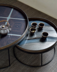 Two round nesting tables with dark metal legs and patterned glass tops, styled with a small teapot and four cups on a textured gray floor.