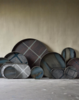 A collection of round decorative trays with various geometric and abstract patterns in shades of blue, gray, brown, and white, arranged against a textured beige wall and concrete floor.