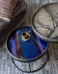 Two round nesting side tables on a cobblestone floor next to a dark upholstered chair. The larger table has a light wood top with a black tree branch pattern, and the smaller table has a glossy blue and purple iridescent glass top with a small cup of coffee on it.