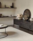 A dark carved wooden sideboard in a styled living room setting, decorated with a round tree art print, a stone sculpture, and a black pedestal lamp, next to a round coffee table on a light rug.