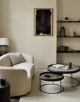 A modern living room scene featuring a cream-colored curved armchair with a textured throw pillow, nested round coffee tables with dark stone tops and black metal frames, a ribbed black side table with a cup, a floor lamp, and a framed abstract print on a beige wall.
