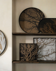 A set of wall art pieces featuring tree branch designs in black, brown, and white, displayed on dark wooden shelves against a warm beige wall.