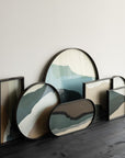 A set of modern abstract landscape wall art panels cum serving tray in various shapes with black frames, leaning against a light gray wall on a dark wood floor.