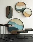 Modern wall decor set featuring four abstract landscape art pieces in muted beige, blue, green, and brown tones, styled in a minimalist room with matching nesting side tables and a glass-top tray on a dark wood floor.