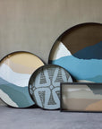 A set of round and rectangular decorative wall art pieces with abstract mountain-like designs in blue, beige, white, and brown tones, displayed against a neutral concrete wall.