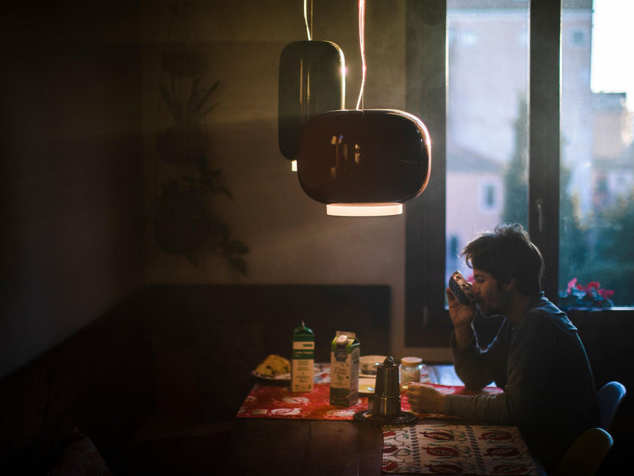 Person sitting at a table in a dimly lit room with a window in the background and a round and long suspension light from Foscarini hanging from the ceiling. 