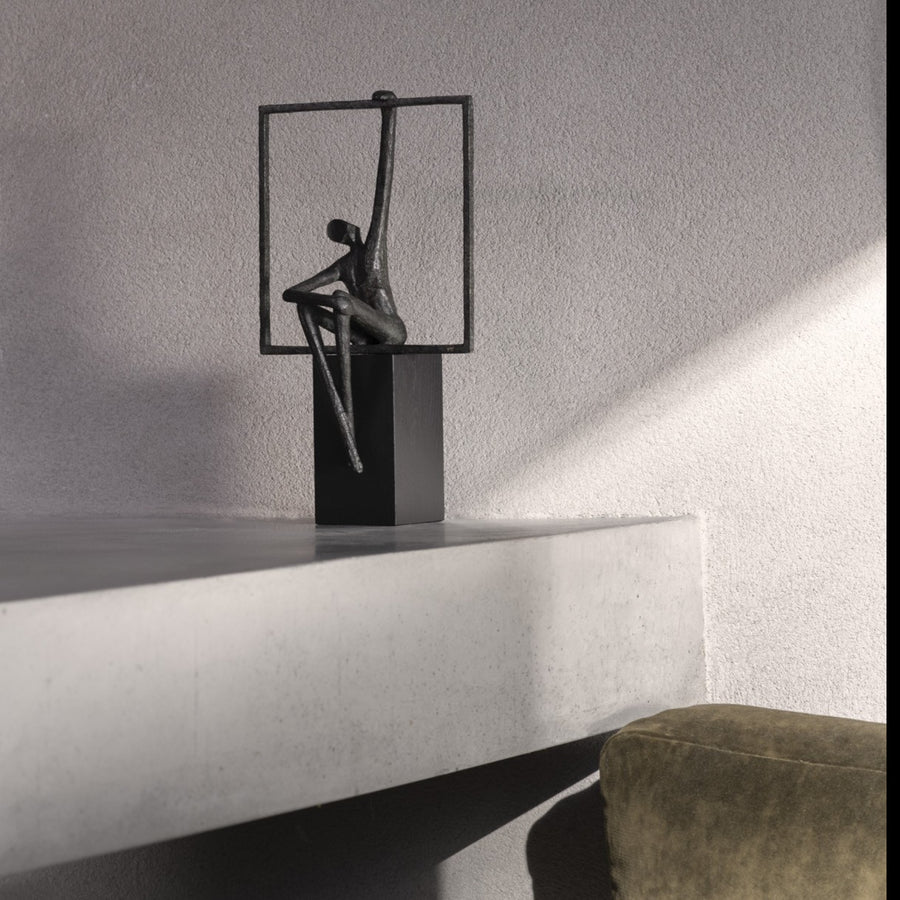 Sculpture of a person sitting on a square platform against a gray wall.