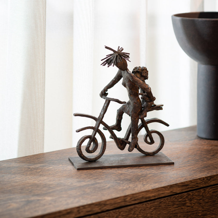 Bronze sculpture of a person on a bicycle with an umbrella on a wooden surface.
