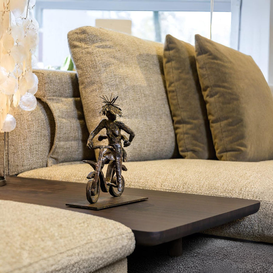 Bronze sculpture of a person on a bike on a coffee table in front of a beige sofa with green pillows.