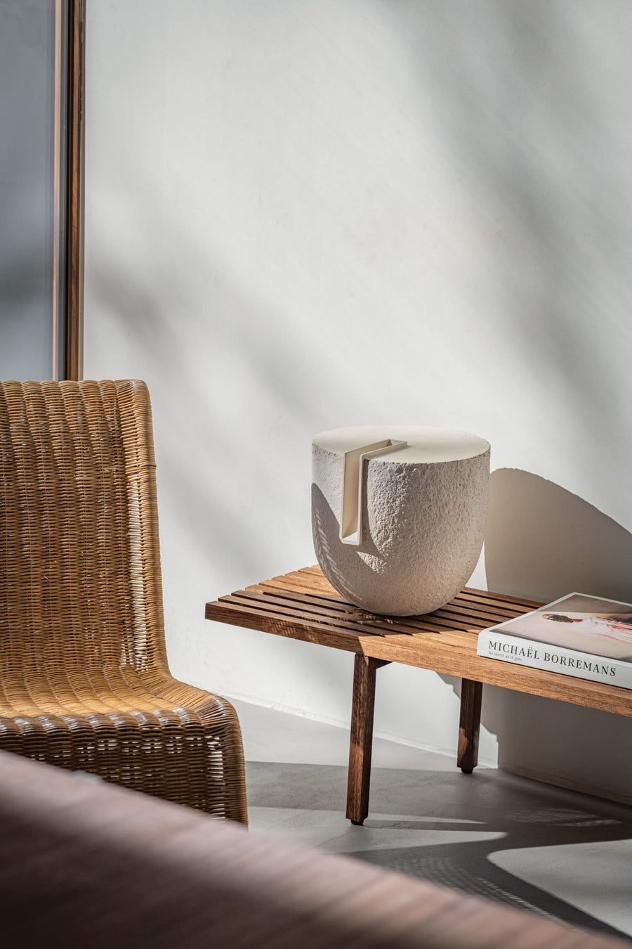 Wicker chair next to a wooden side table with a decorative sculpture from Gardeco and book against a white wall.