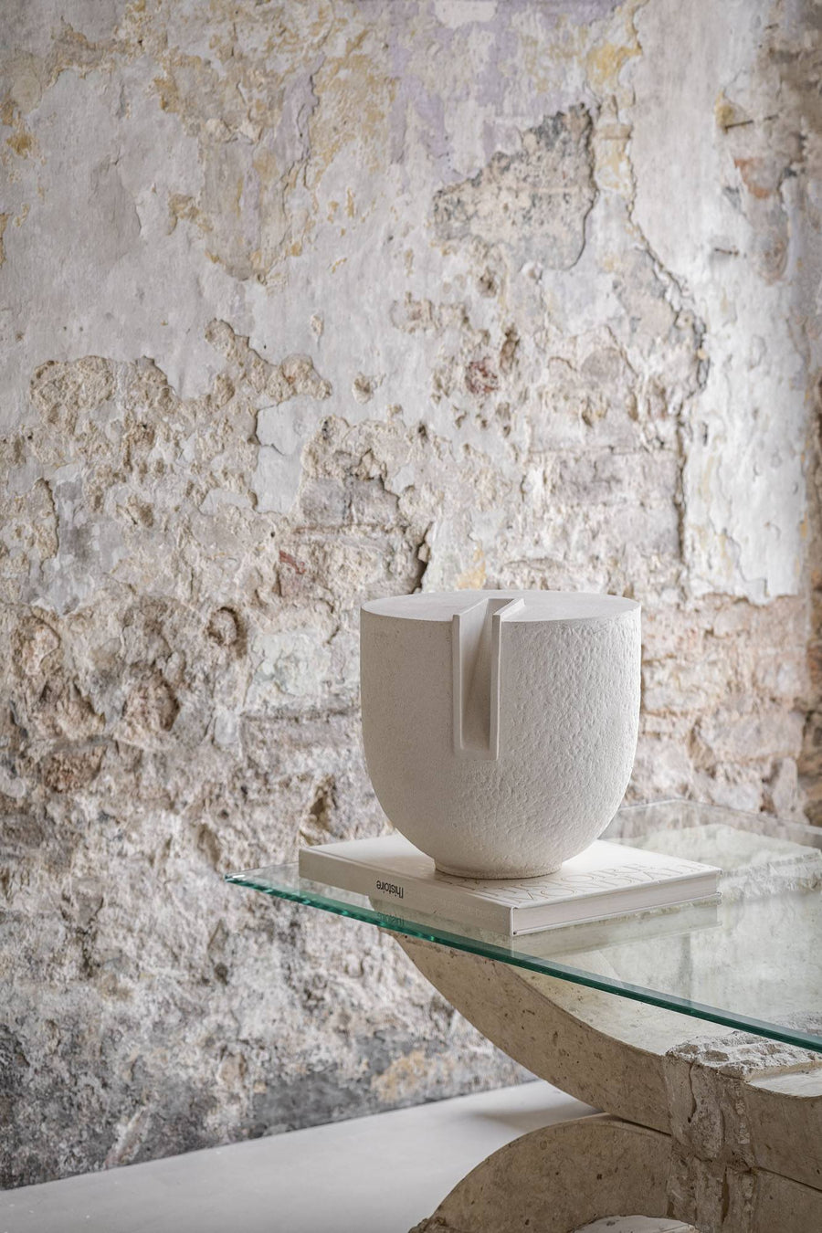 White ceramic sculpture on a glass table against a textured stone wall