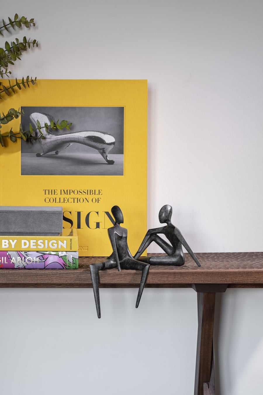 Book titled 'The Impossible Collection of Design' by Assouline on a shelf with decorative bronze statues and plants.