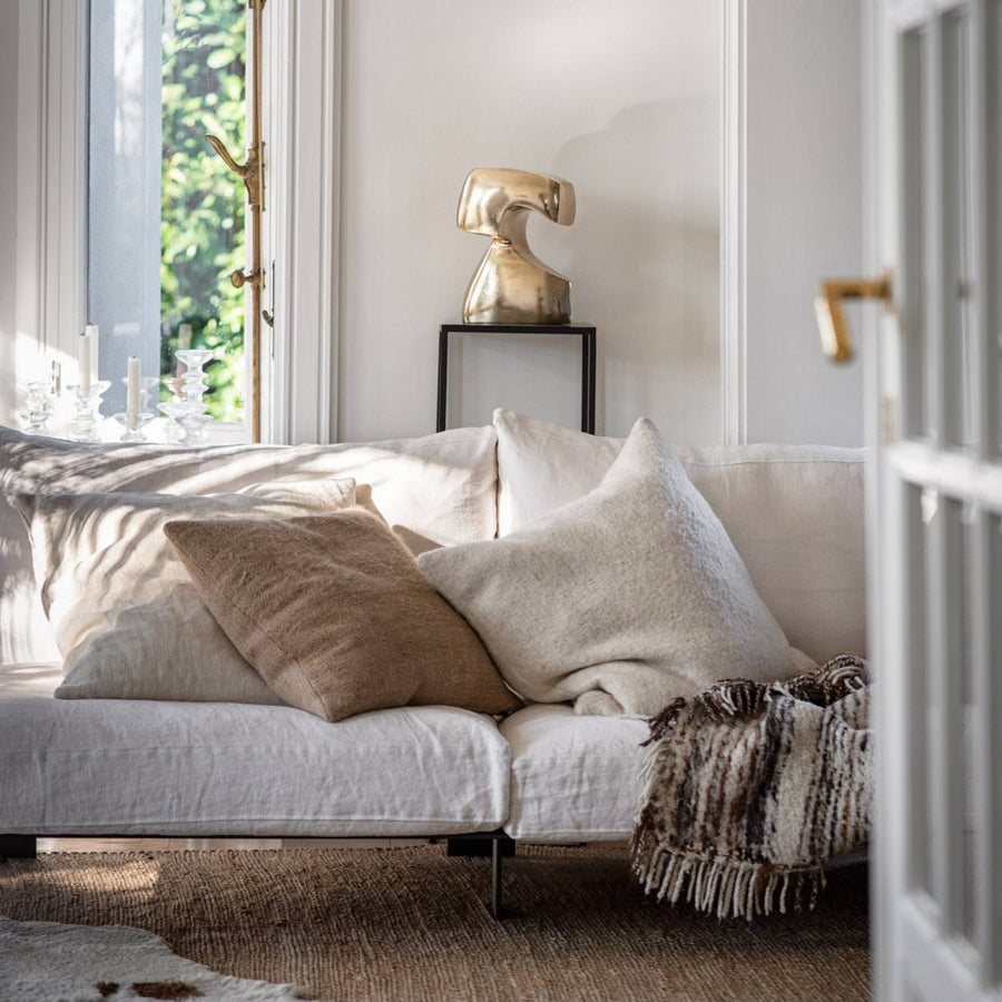 White sofa with beige and brown cushions in a bright room with natural light and a polished bronze sculpture on a high pedestal. 