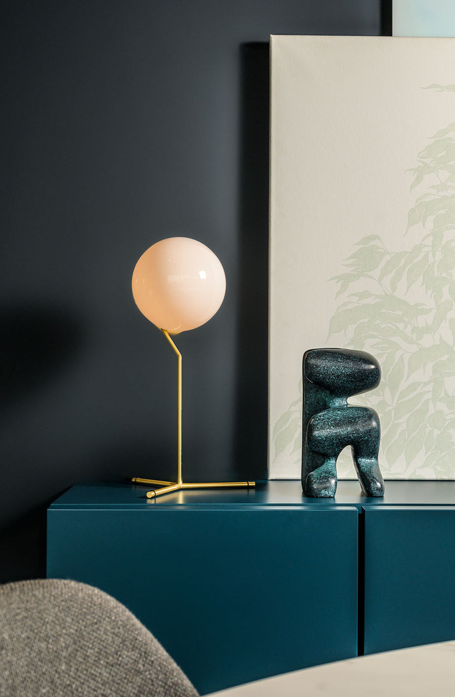 Modern interior with a table lamp and decorative object on a dark blue cabinet against a dark wall.