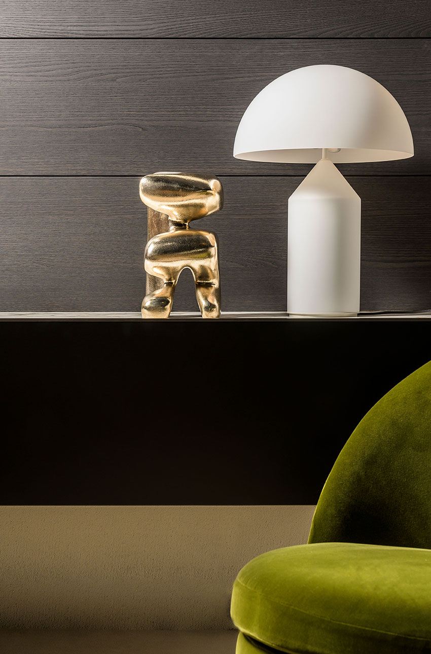 White lamp and polished bronze sculpture on a dark surface with a green chair in the foreground.