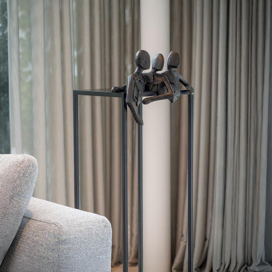 Decorative sculpture of three figures on a stand in a living room setting with curtains and a sofa.