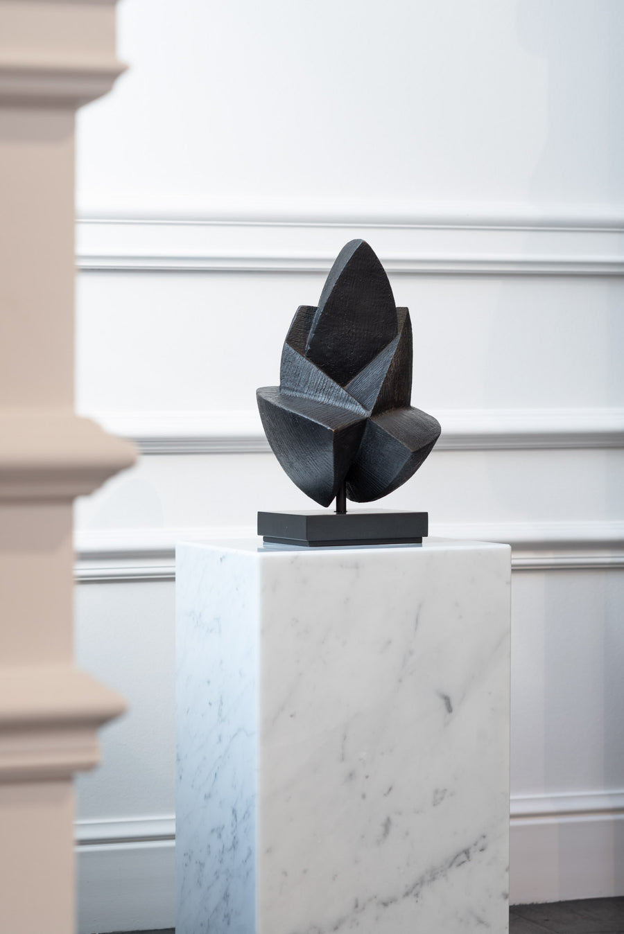 Abstract sculpture on a marble pedestal against a white wall.