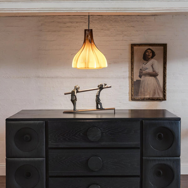 A bronze sculpture on a wooden sideboard below a yellow glass lamp and next to a lady portrait on a textured wall