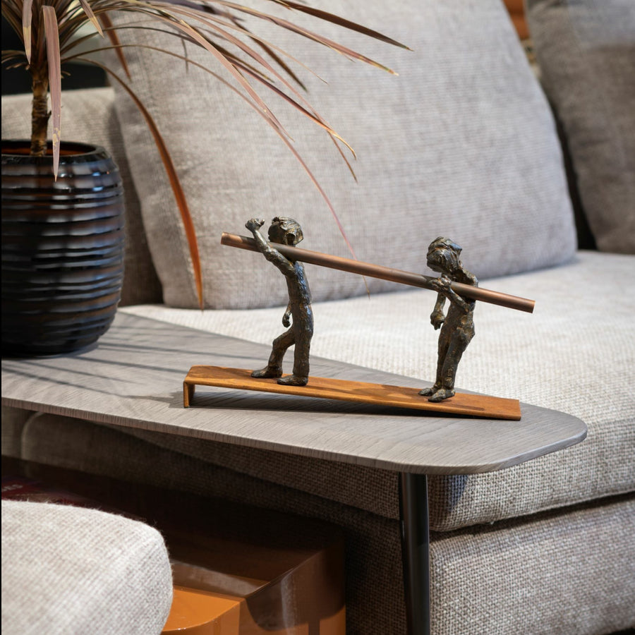 Bronze sculpture of two figures carrying a log on a wooden base, placed on a coffee table.