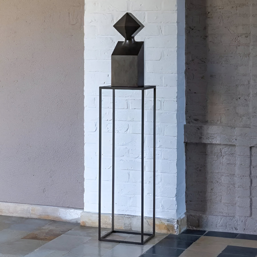 Black geometric bronze sculpture on a tall black stand against a white brick wall.