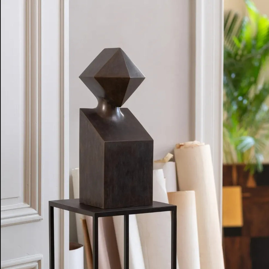 Abstract black metal sculpture on a stand against a neutral wall with a plant in the background
