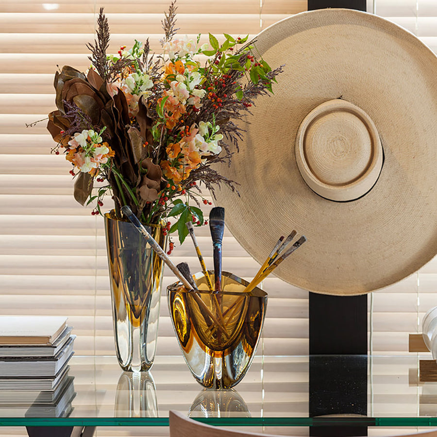 An exquisite Gardeco Glass Vase Triangle 4 Fume Amber filled with elegant flowers and delicate brushes, showcased among other glass objects.