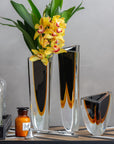 A Gardeco Glass Vase Triangle 1 Black Amber with flowers and a bottle of alcohol featuring contemporary refinement and bicolour glass.