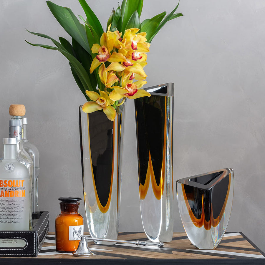 A Gardeco Glass Vase Triangle 1 Black Amber with flowers and a bottle of alcohol featuring contemporary refinement and bicolour glass.