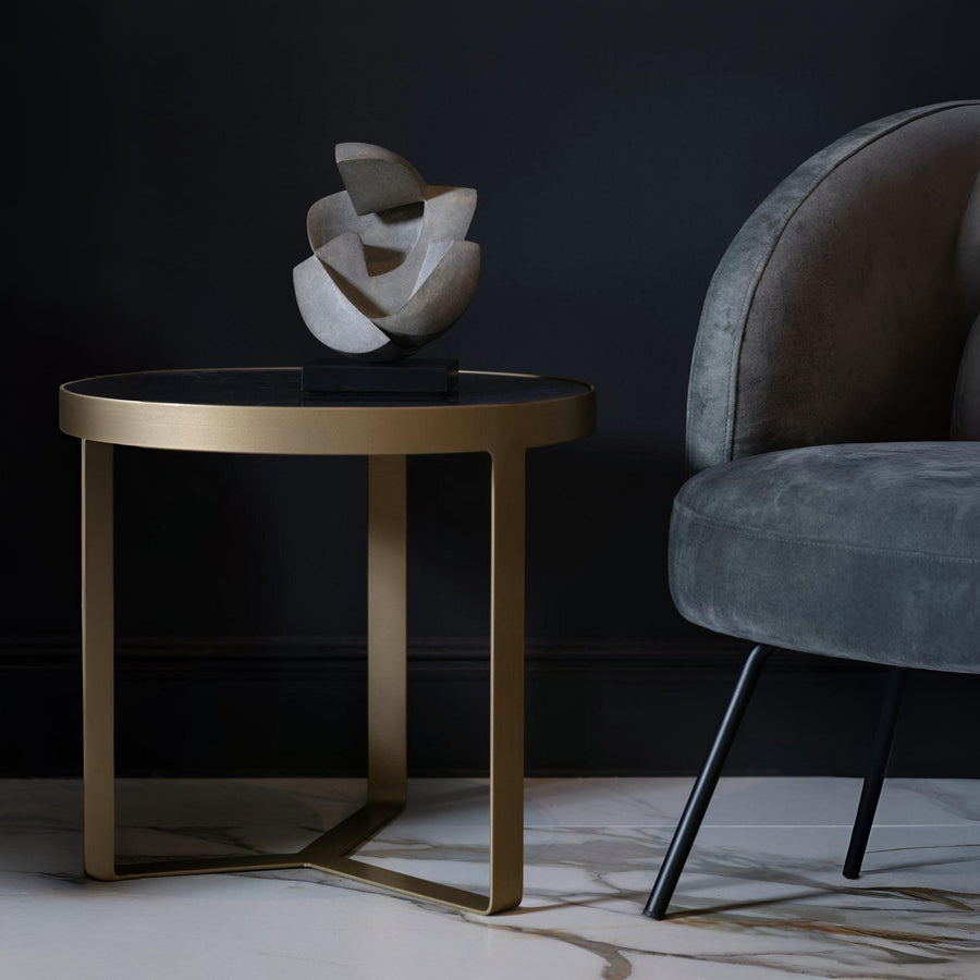 Abstract sculpture on a round table next to a gray armchair against a dark wall.