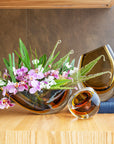 A Gardeco Glass Vase 96 Fume Amber with Gardeco artisanal craftsmanship, adorned with beautiful flowers and a delicate glass ball, placed elegantly on a table.