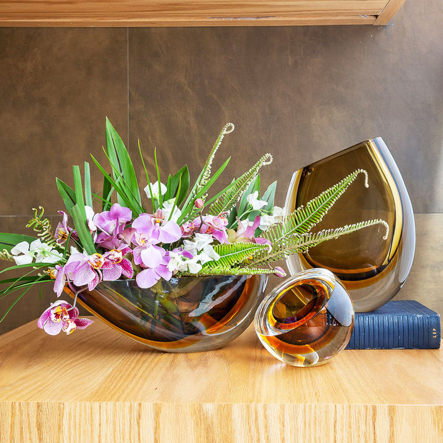 A Gardeco Glass Vase 96 Fume Amber with Gardeco artisanal craftsmanship, adorned with beautiful flowers and a delicate glass ball, placed elegantly on a table.