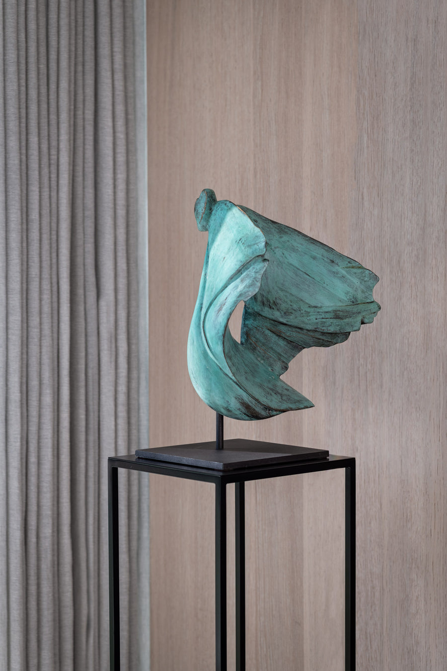 Teal abstract sculpture on a black stand against a wooden paneled wall.