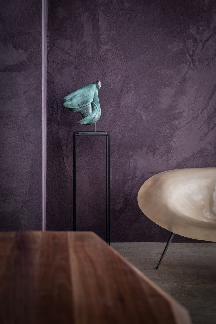 Bronze sculpture on a stand against a textured purple wall with a wooden table and chair in the foreground.