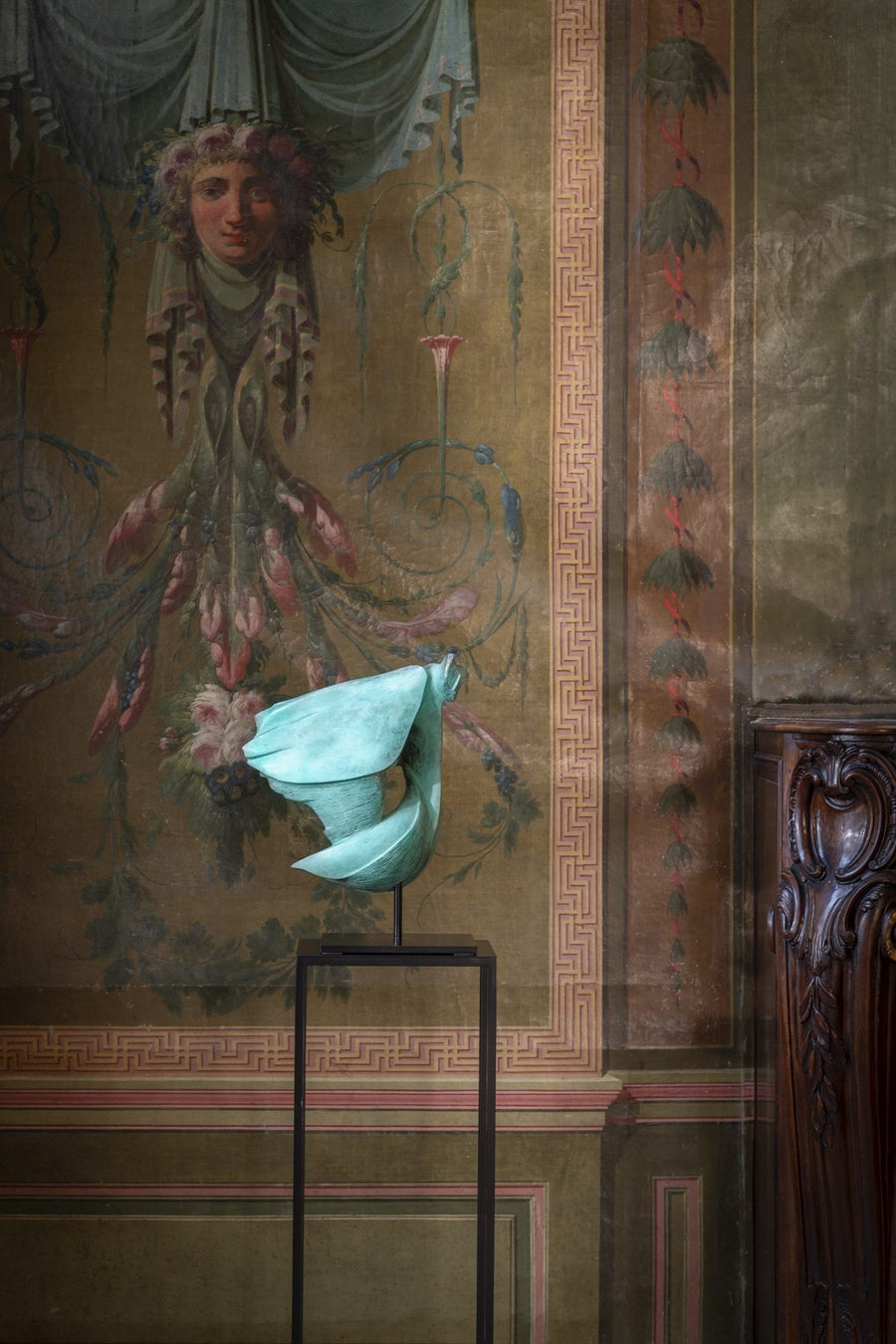 Decorative wall with classical painting and a teal flower on a stand.