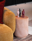 Round pink side table with decorative figurines next to a mustard yellow armchair with a red cushion.