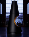 A modern black cone-shaped table or lamp with a crescent cutout on the side, emitting a warm orange glow from within, set in a dark, atmospheric industrial interior.