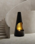 A modern black freestanding bioethanol fireplace with a curved gold-toned firebox and visible flames, placed in a minimalist interior with beige walls and a curved staircase.