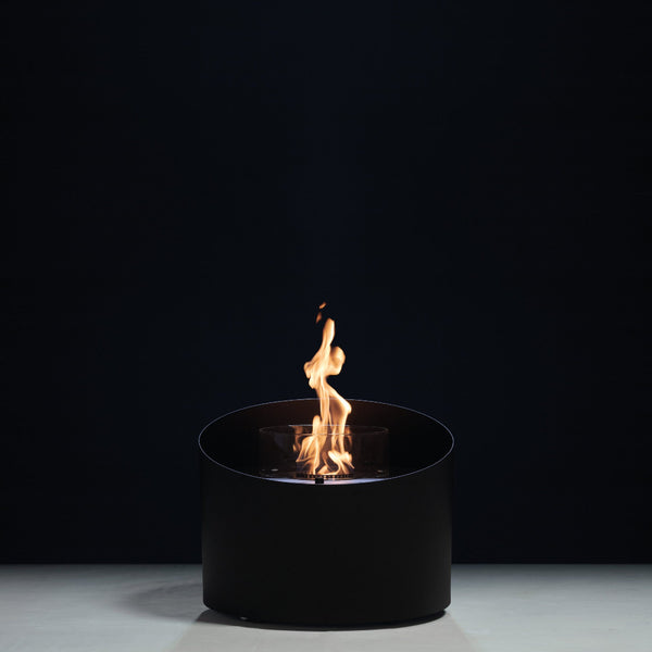 A modern black cylindrical bioethanol fireplace with a visible orange and yellow flame, set against a dark studio background.