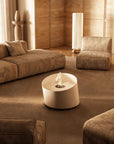 A modern cylindrical white coffee table with a small flame feature in the center, set in a warm, contemporary living room with beige sofas and wood-paneled walls.