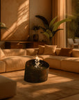 A modern sectional sofa in warm tan and brown tones arranged in a sunlit living room with a round bioethanol fireplace, large plants, and floor-to-ceiling windows.