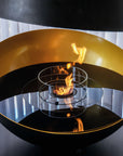 A modern black and gold metal fire bowl with a glass wind guard and active flame, set against a background of white vertical blinds.