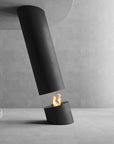 A modern black cylindrical tabletop fireplace with an open top and visible flame, set against a minimalist concrete wall and floor.