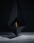 A modern black geometric fire pit with a suspended triangular design, featuring a small gold flame element in the center, set against a dark studio background.