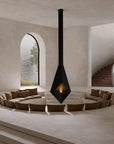 A modern black cone-shaped indoor fireplace with a visible flame, suspended over a circular beige lounge seating arrangement in a minimalist, warm-toned stone interior with arched windows and a staircase.