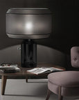 Modern table lamp on a wooden side table with a chair and books in a dimly lit room.