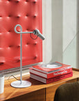 White desk lamp on a wooden surface with books and a mug in front of a red textured wall.