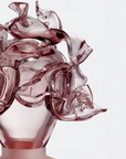 A close-up of a sculptural object or vase with flowing, translucent burgundy-pink glass on a clear base against a light gray background.