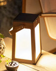 Wooden lantern-style lamp emitting a warm glow on a wooden surface with a plant and decorative item in the background.
