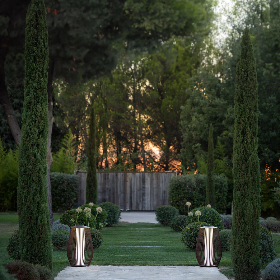 Garden pathway with tall green trees and decorative solar lanterns at dusk by Les Jardins available at Spacio retail stores in Mumbai & Kolkata. 